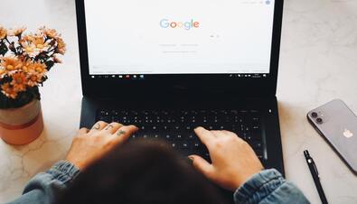 picture is a view of someone's hands on a laptop which is open to the Google search page. there is an iphone and a pen to their right and flowers in a pot to their left. 